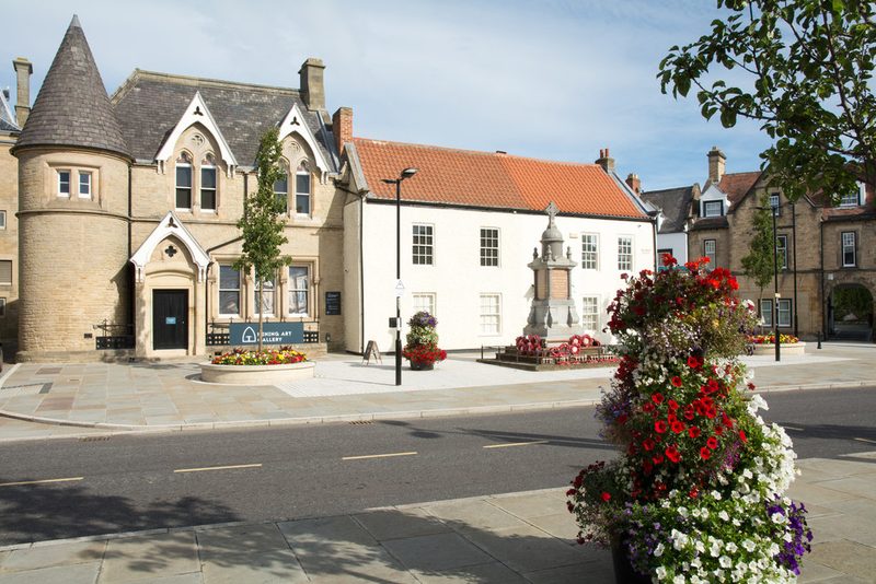 Stone building with a round corner turret and a Mining Art Gallery sign, seen across Bishop Auckland Market Place