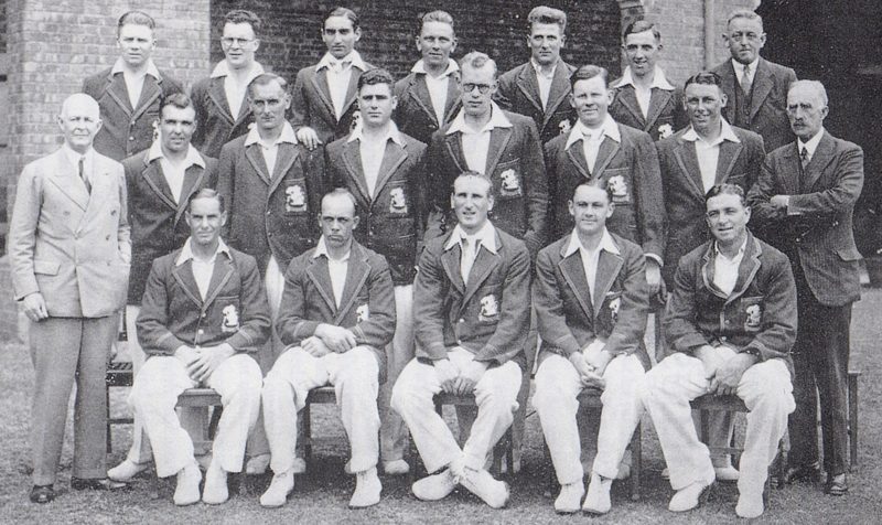 Black and white team photograph of three rows of men in dark blazers and white trousers in front of a brick wall, two officials in suits at the ends.