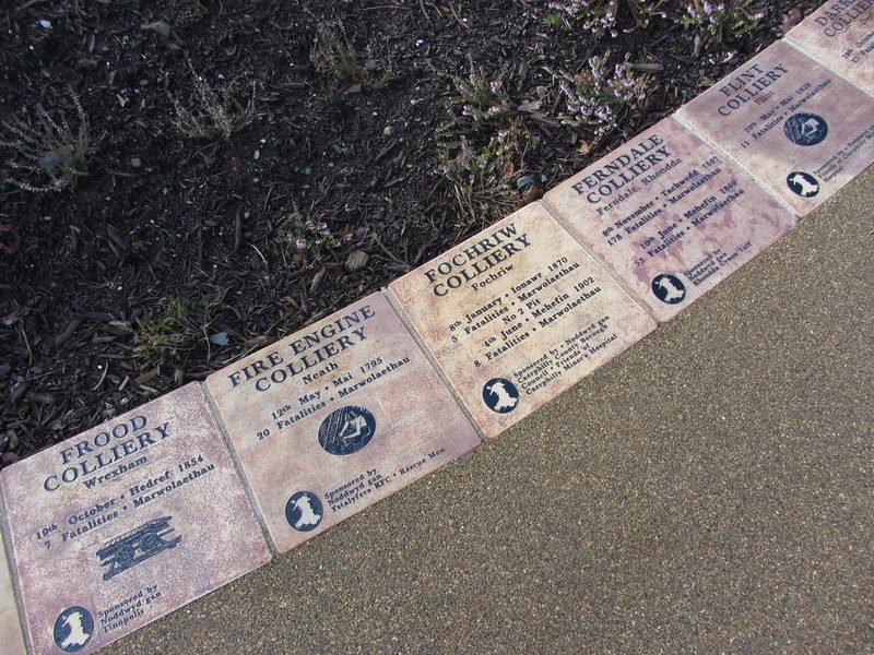 A row of square ceramic tiles set in paving, each naming a Welsh colliery, its disaster date and its dead.