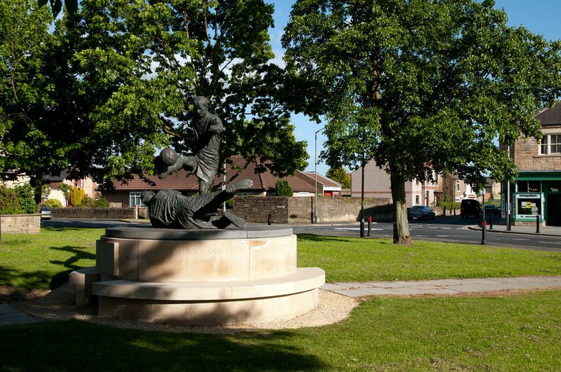 Bronze statue of two figures, one heading a football, on a round stone plinth on a village green.