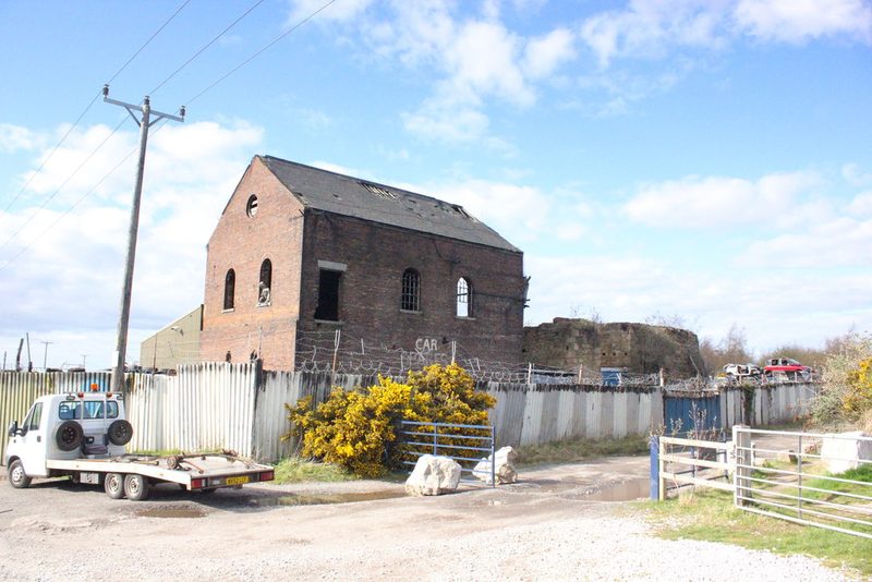 A roofless red-brick engine house stands behind a corrugated fence and a gorse bush beside a gravel track.