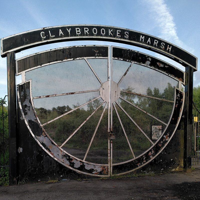 Black metal gates shaped like a half pit wheel stand beneath an arched sign reading Claybrookes Marsh.