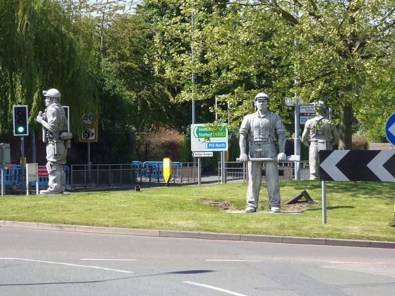 Bronze sculptures of miners standing on a paved traffic island in a town centre, with shrub planting around the base and buildings behind.