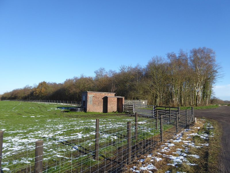 A small brick building beside a fenced field and woodland, with patches of snow on the grass.