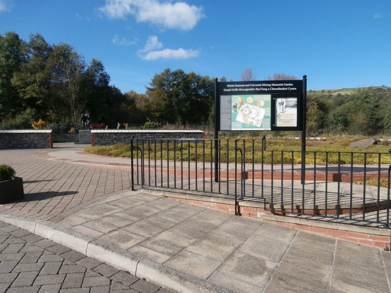 Paved approach to a memorial garden with a railing, an information board and low stone walls under a blue sky.