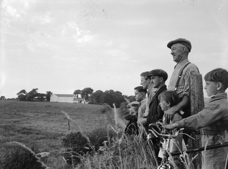 A row of men and children in caps stand by a fence looking across a field towards farm buildings.