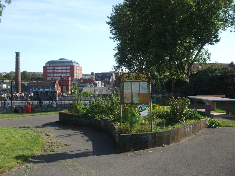 A community garden bed and noticeboard in a park, with a tall brick chimney beyond the railings.