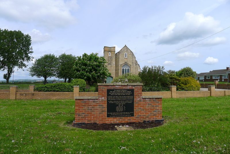 A brick memorial wall with a black inscribed plaque on a grass verge, with a stone church behind.