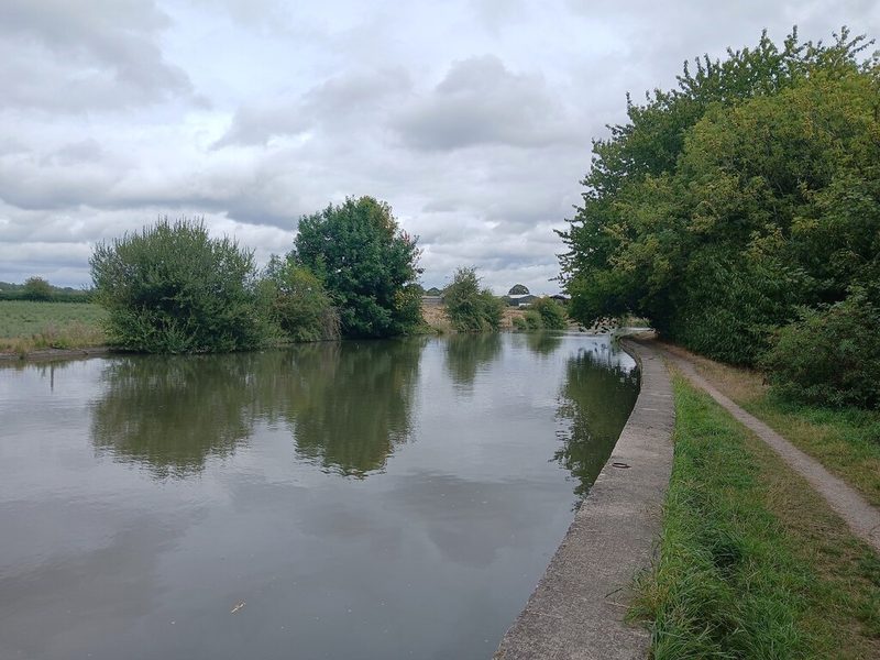 A still, wide canal with a concrete-edged towpath on the right and trees on both banks under grey cloud.