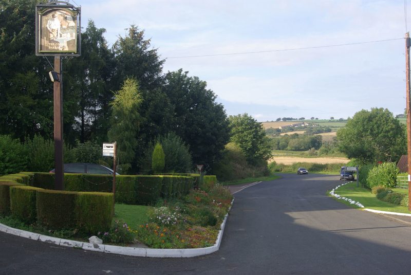 A village road curving past a hedge and flower bed, with a hanging inn sign on a post and fields beyond.