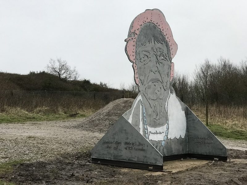 A painted steel cut-out of a miner in a cap on an inscribed base, on the grassed colliery site.