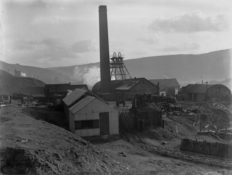 A colliery headframe and a tall chimney rise behind sheds and stacked pit props in a hillside valley.