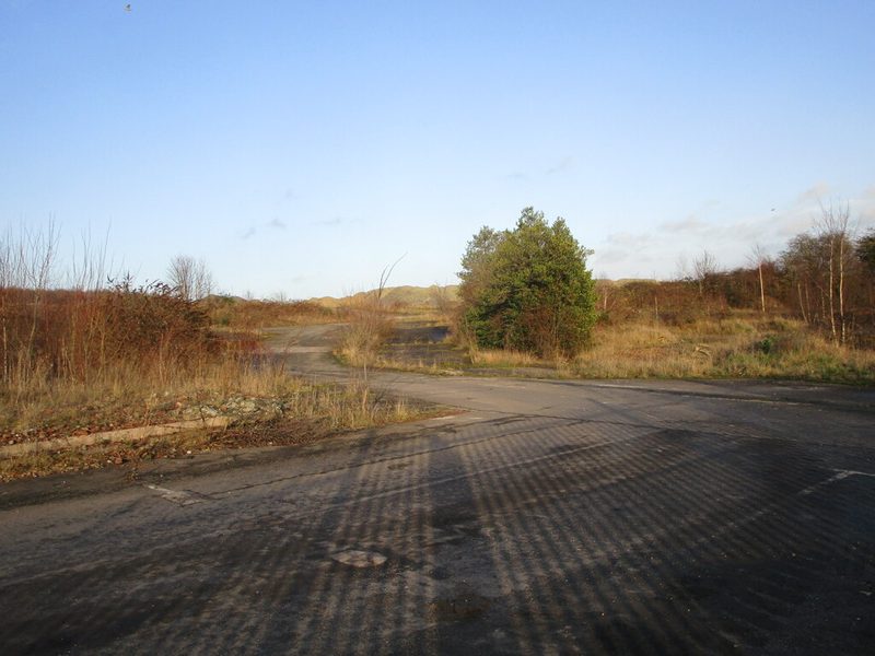 An empty area of cracked tarmac and rough ground where a colliery once stood, under a blue sky.