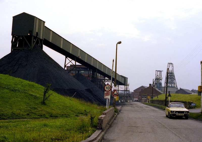 A colliery in 1979 with headgear, a raised conveyor and a large stack of coal beside a road.