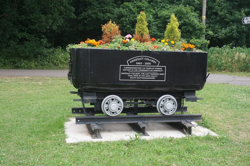 A stone memorial to Annesley Colliery standing on mown grass, with a pit winding wheel set behind it.