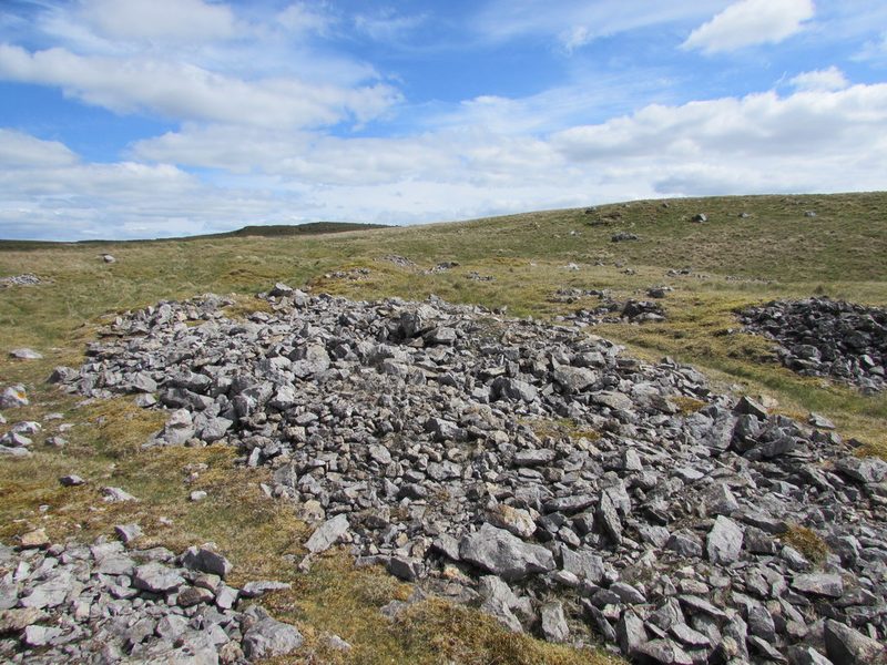 A heap of grey limestone spoil on open moorland, with further hollows and stone scatters on the slope behind under a blue sky.