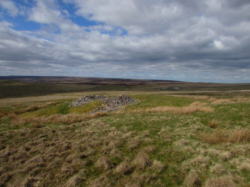 Two low mounds of grey stone spoil beside a filled shaft on rough moorland grass, with brown moors stretching to the horizon under cloud.