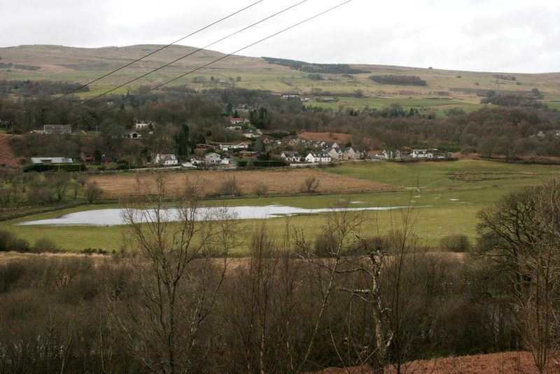 Flooded fields in a valley below wooded rising ground at the edge of a town.
