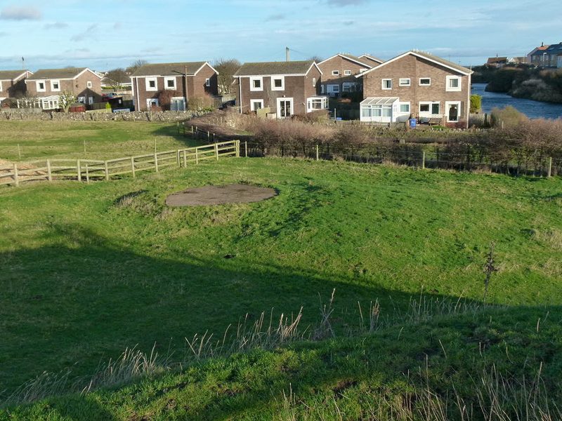 A round concrete cap over the colliery shaft in a grassy field, with houses and a flooded quarry behind.