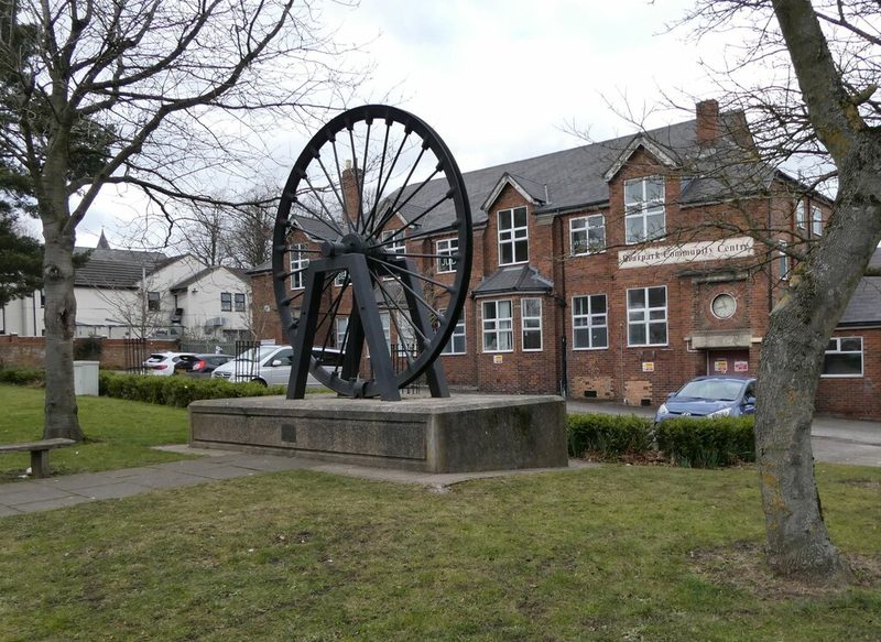 A pit wheel on a stone plinth on a green in front of a two-storey red brick building signed Bearpark Community Centre.