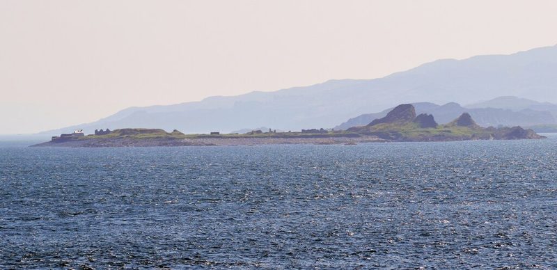 A low island seen across open water, with the roofless walls of quarry buildings along its length and Scarba's hills behind.