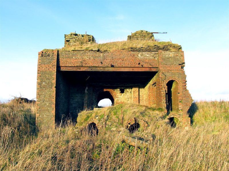 The ruined stone and brick engine house at Caer Eithin standing on open hilltop ground above Three Crosses, with the mouth of its vaulted tunnel visible.