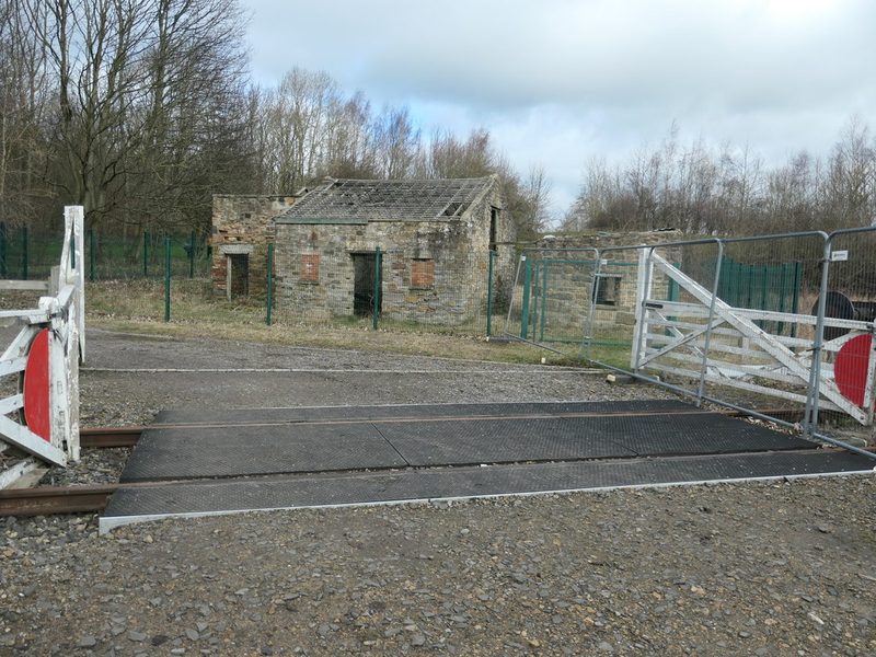 A roofless stone building behind green fencing beside a gated level crossing on an old railway line.