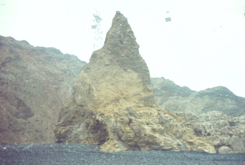 A stone pinnacle rising from a beach black with coal waste at Blackhall, with the colliery's overhead bucket ropeway running out above the shore.