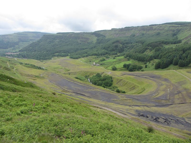 A wide grassed-over colliery site with bare black ground curving round a wooded stream below forested valley sides.