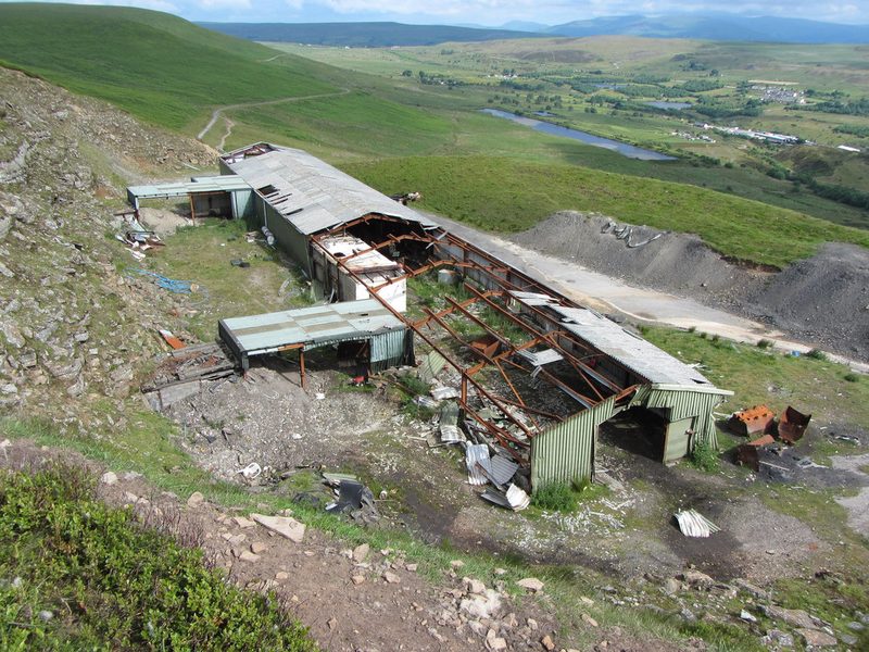 A large derelict industrial shed cut into a hillside, with drift entrances and old drams nearby and lakes in the distance.