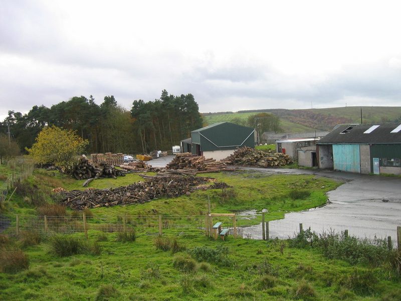 Sheds and a yard stacked with timber and logs below a conifer plantation on the former drift site