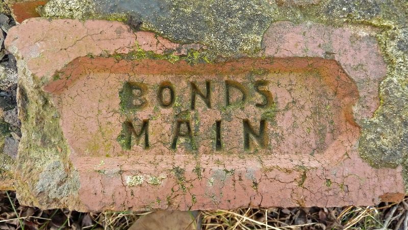 A weathered red brick with the words BONDS MAIN pressed into its frog, lying among stones and dry grass.