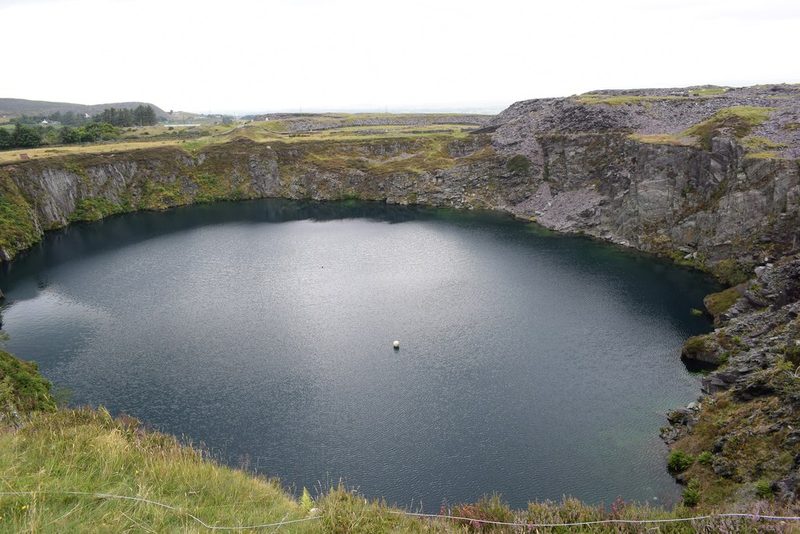 A large round pool of dark water fills a quarry pit ringed by grey rock faces and slate waste under a pale sky.