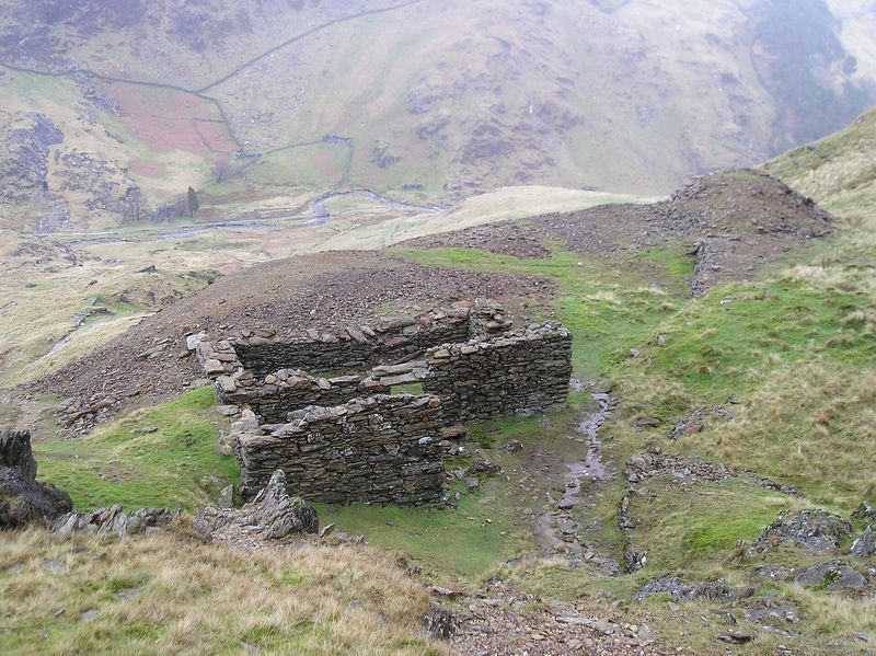 A roofless drystone building beside a grey spoil heap on a steep hillside, with a valley and stream far below.