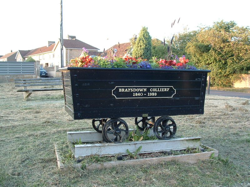 A wooden and iron mine tub standing on a paved area in Peasedown St John, filled with earth and planted as a flower bed.