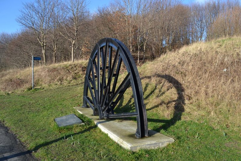 A black cast-iron pit wheel set upright on a concrete plinth beside a grassy bank, with a stone plaque in front.
