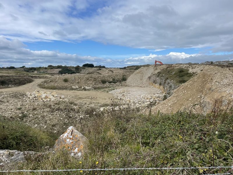 A working limestone quarry with heaps of cut blocks and rubble, an orange excavator on the far rim and houses beyond.