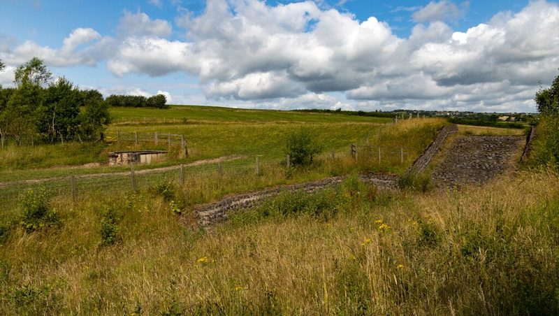 Rough grassland with stone-lined drainage channels and a small concrete structure under a cloudy sky.