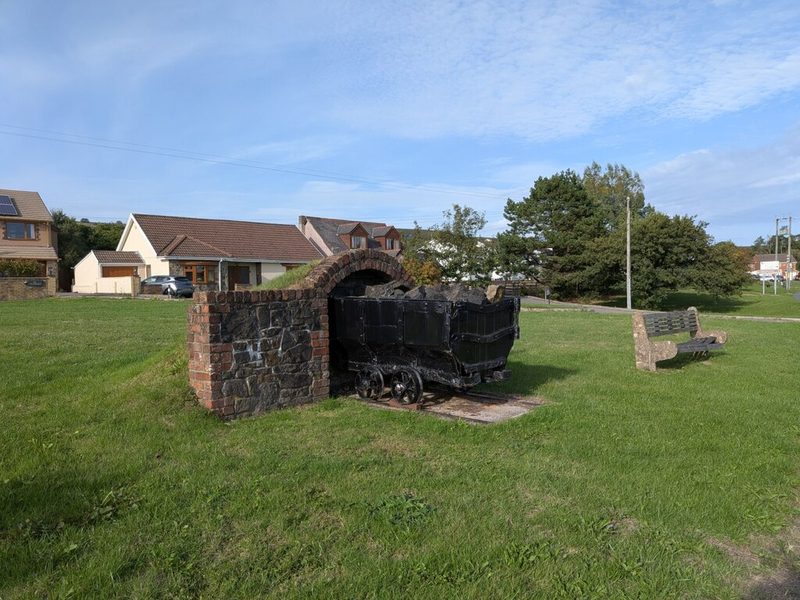 A full coal dram on rails entering a stone-built tunnel mouth, set on a grassy bank as a miners' memorial.