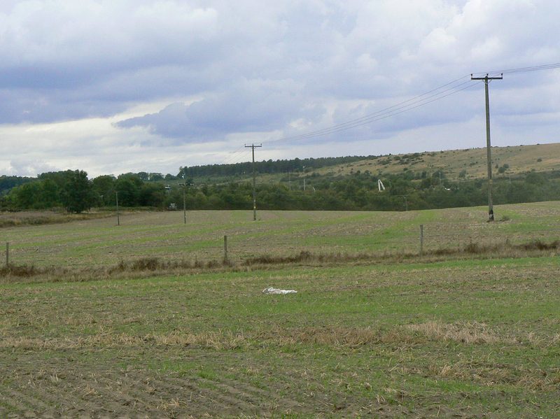 Arable fields crossed by power lines, leading to a belt of trees and a grassy hillside behind which lies the cleared colliery site