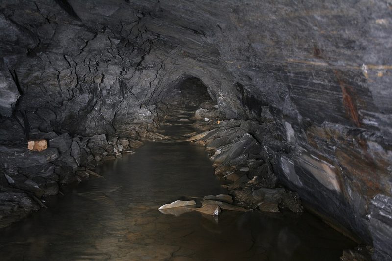 A low arched tunnel cut through dark grey slate, its floor covered by still water with slabs of slate along the edges.