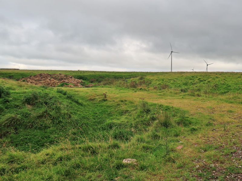 Rough grassy ground with hollows and a heap of earth, under a grey sky with wind turbines on the horizon.