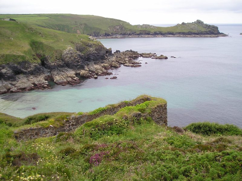 A ruined stone wheelpit on a grassy clifftop above a rocky cove, with a headland and the sea beyond.