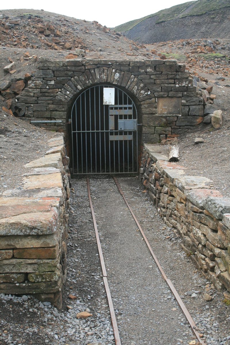 A gated stone archway set into a hillside, the entrance to a mine level with a track leading in