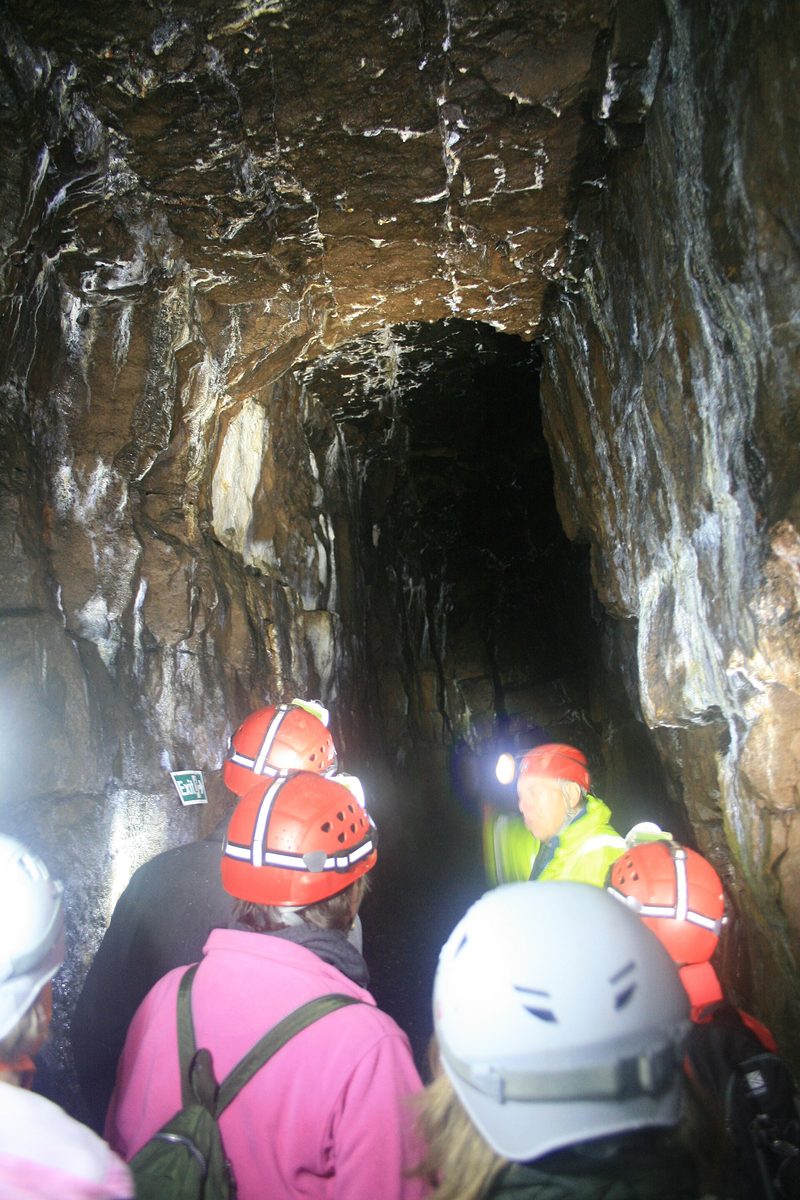 A visitor walking along a narrow stone-lined mine level lit by lamps, with rails on the floor