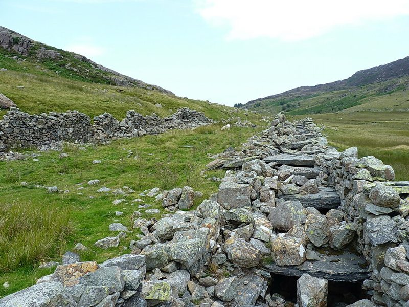 Ruined drystone walls of grey slate blocks run along a grassy valley floor between rocky hillsides.