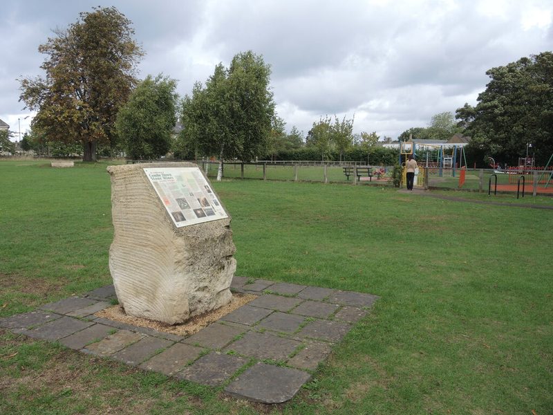 A rough block of Bath stone carrying an information panel about the stone mines, standing on a paved square in a grassy park with a playground behind.