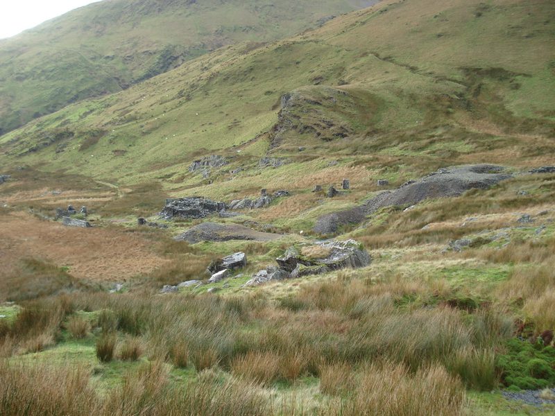 Scattered stone ruins, pillars and grey spoil heaps lie on a rough grassy valley floor below green hillsides.