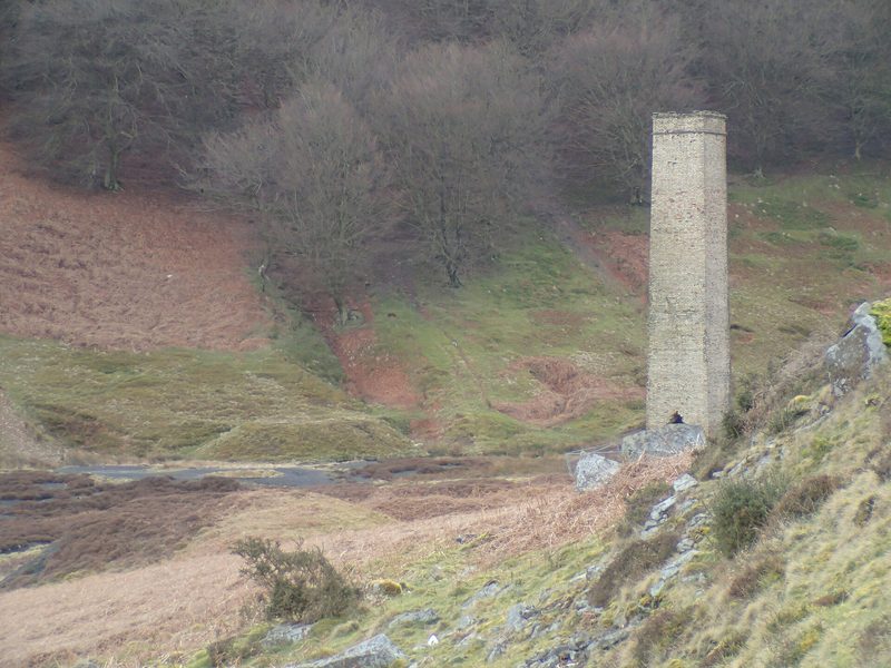 A tall slender brick chimney standing alone on a grassy hillside above a stream, with old spoil mounds around it and bare woodland behind.