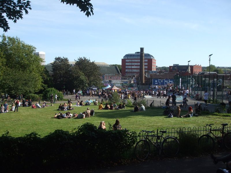 Crowds on the grass of a city park on a sunny day, with a tall brick chimney and red-brick buildings beyond.